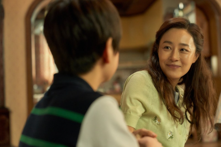한 여성이 미소를 지으며 앞의 소년을 바라보고 있다. 여성은 연두색 스웨터를 입고 있으며, 따뜻하고 인자한 표정으로 소년과 마주 앉아 있다. 소년의 뒷모습이 보이고, 두 사람의 모습에서 다정한 분위기가 느껴진다.