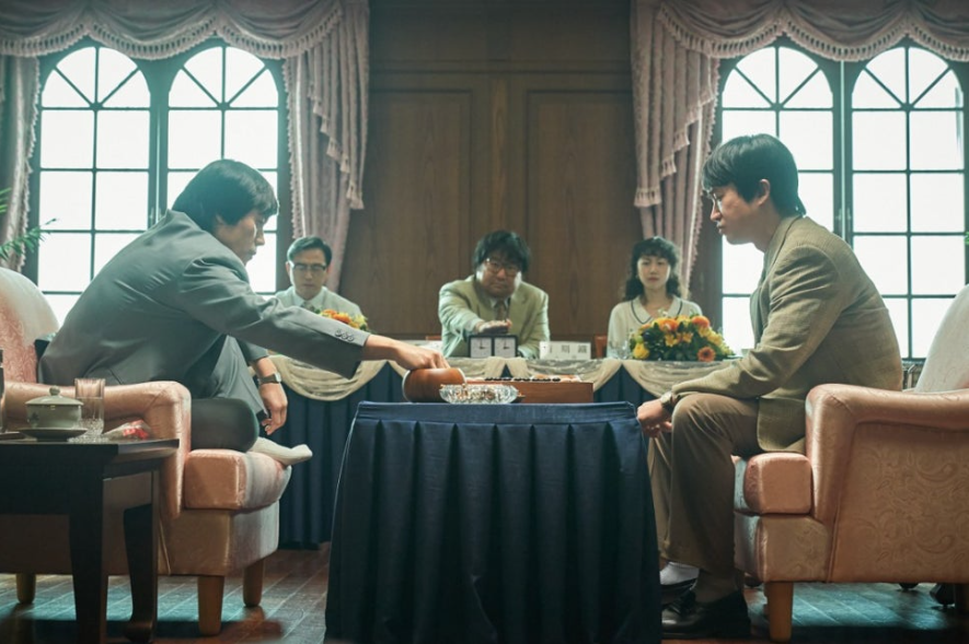 두 남성이 테이블에 앉아 바둑을 두고 있다. 한 남성은 바둑돌을 내려놓기 위해 몸을 숙이고 있고, 다른 남성은 진지한 표정으로 이를 지켜본다. 주변에는 다른 사람들이 앉아 경기를 관전하고 있다.