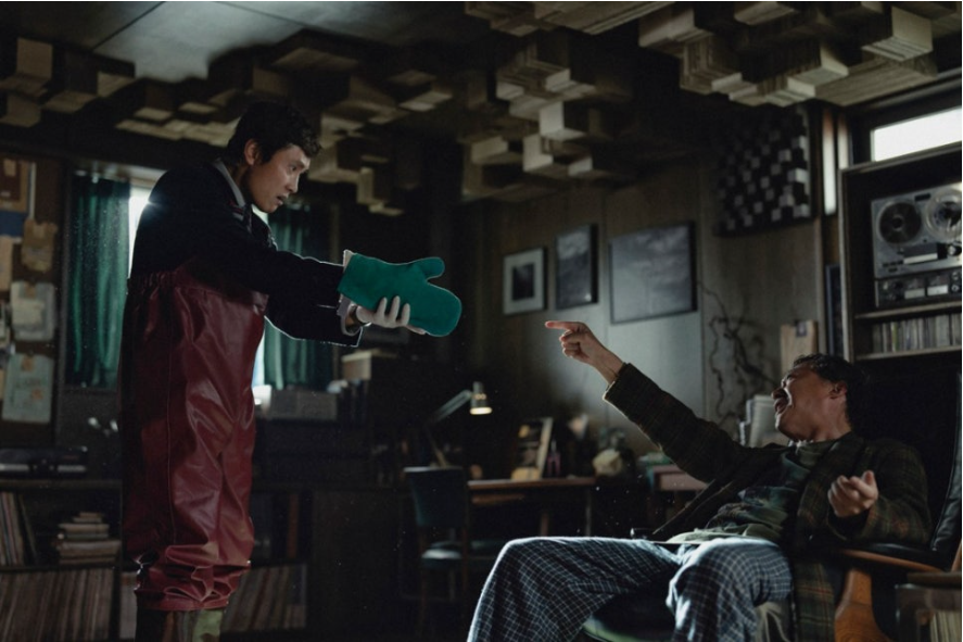 방수 바지를 입은 만수가 한 남성에게 무언가를 건네고 있고, 남성은 의자에 기대 앉아 만수를 손가락으로 가리키며 화를 내는 듯합니다. 독특한 인테리어의 방에서 심각한 상황이 연출됩니다.