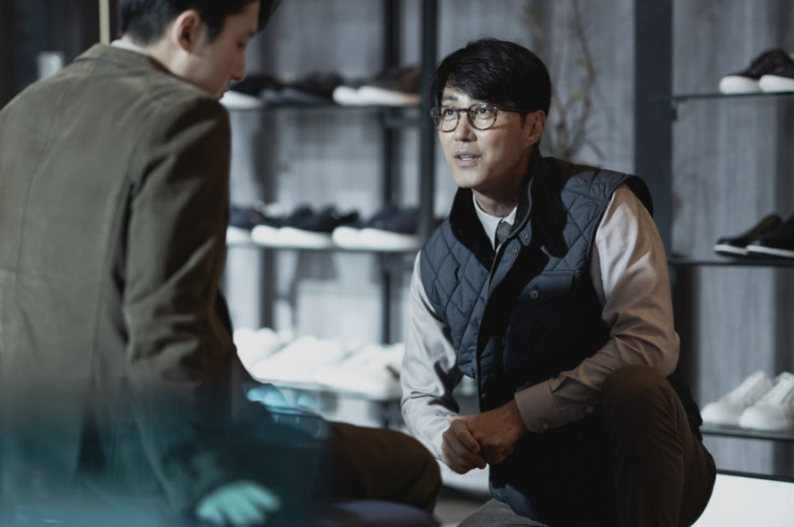 고시조가 안경을 쓰고 고객에게 웅크려 앉아 친절하게 구두를 설명하는 듯한 모습에서, 과거 제지업계 기술자였던 그의 변화된 상황이 드러납니다.