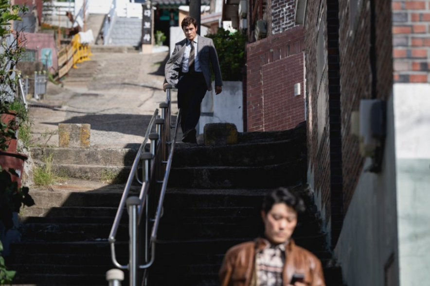 이 스틸컷은 영화 어쩔 수가 없다에서 주인공 유만수(이병헌)가 언덕길의 계단을 내려오는 모습입니다. 만수는 흐트러진 정장 차림으로 지쳐 보이는데, 아래쪽에는 최선출(박희순) 가죽 재킷을 입고 핸드폰을 보고 있습니다. 두 사람의 엇갈린 처지와 긴장감이 느껴집니다.