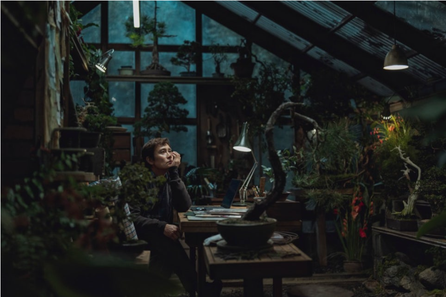 유만수(이병헌)가 어두운 공간에 앉아 있습니다. 긴장한 표정과 단정한 모습에서 재취업을 향한 압박감이 느껴집니다.