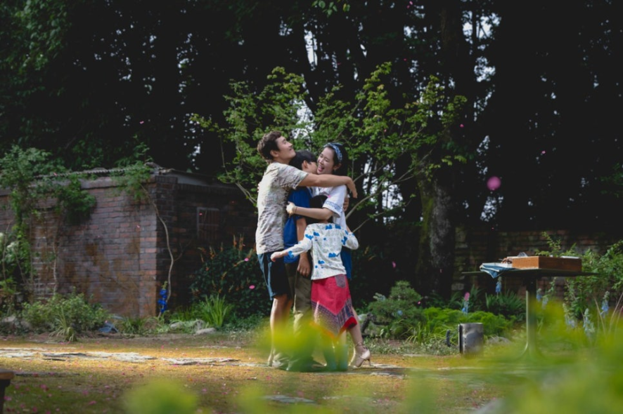 주인공 유만수 가족이 햇살 좋은 뜰에서 서로를 꼭 껴안고 환하게 웃고 있습니다. 만수(이병헌)와 미리(손예진) 그리고 두 아이가 화목한 모습으로 가족의 행복한 한때를 보여줍니다.