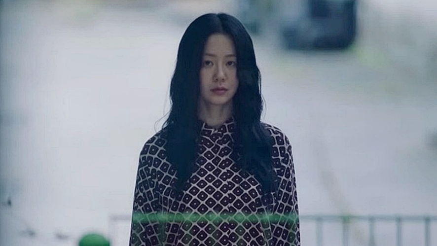 배우 고현정으로 보이는 여주인공이 화면 중앙에 클로즈업되어 있습니다. 그녀는 검은색 긴 생머리를 하고 패턴이 있는 블라우스를 입은 채 어딘가를 응시하고 있습니다. 주변 배경은 다소 흐릿하고 밝아 그녀의 무표정한 얼굴이 더욱 부각됩니다.