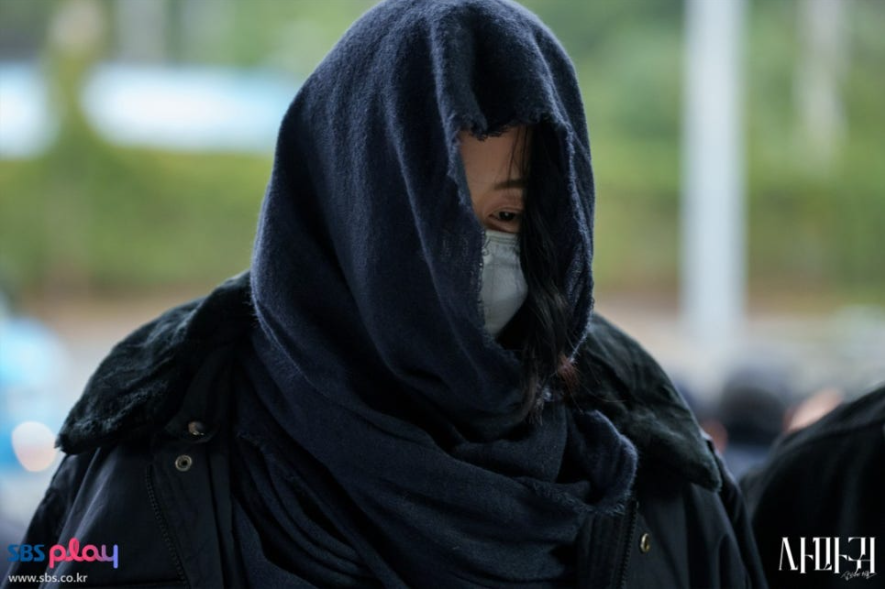 배우 고현정으로 보이는 인물이 어두운 색 스카프나 천으로 머리와 얼굴을 깊게 가리고 마스크를 착용한 모습입니다. 얼굴을 거의 드러내지 않고 고개를 숙인 채 음침하고 비밀스러운 분위기를 풍겨 극 중 캐릭터의 어두운 상황을 짐작하게 합니다.