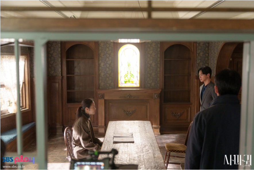 고전적인 분위기의 실내에서 세 인물이 긴 탁자를 사이에 두고 마주하고 있습니다. 고현정으로 보이는 여성 캐릭터는 왼쪽에 앉아 있고, 장동윤으로 보이는 남성 캐릭터는 오른쪽에 서 있으며, 다른 중년 남성이 뒷모습으로 보입니다. 창문의 스테인드글라스가 독특한 분위기를 더합니다.